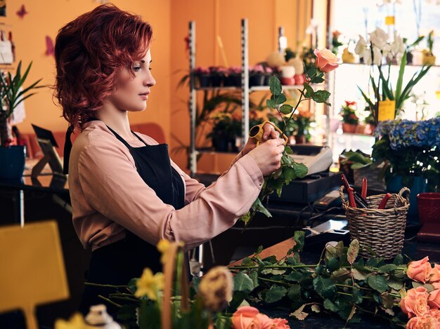Encantadora floristería pelirroja con uniforme haciendo una hermosa composición floral mientras está de pie en el mostrador de una floristería.