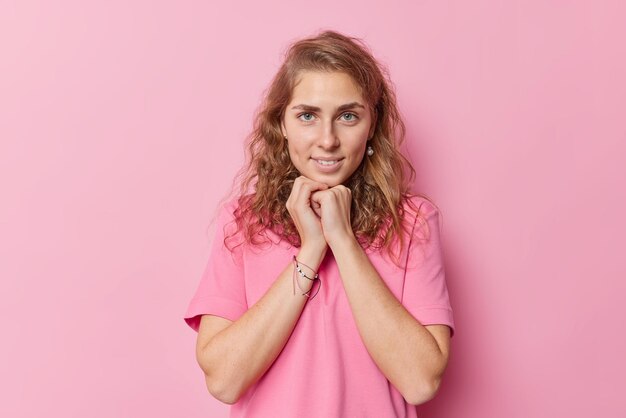 Encantadora y hermosa mujer joven de ojos azules con el pelo largo y ondulado mantiene las manos debajo de la barbilla mira directamente a la cámara vestida con una camiseta básica informal aislada sobre poses de fondo rosa para hacer fotos