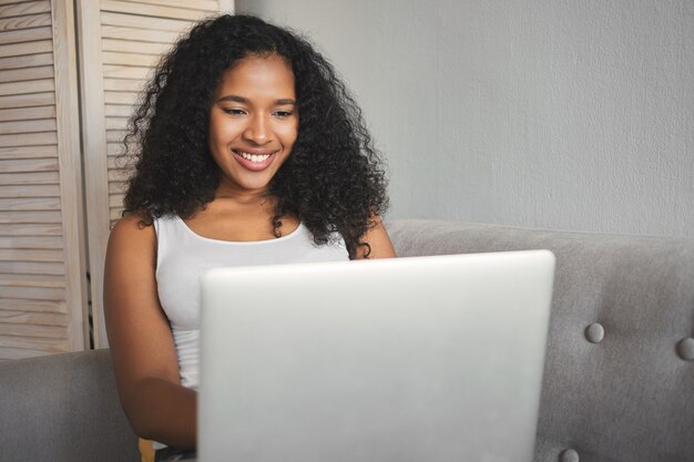 Encantadora joven afroamericana en camiseta blanca sin mangas disfrutando de la comunicación en línea, usando wifi gratis en la computadora portátil, navegando por Internet, viendo videos de blogs o charlando con un amigo, sonriendo felizmente