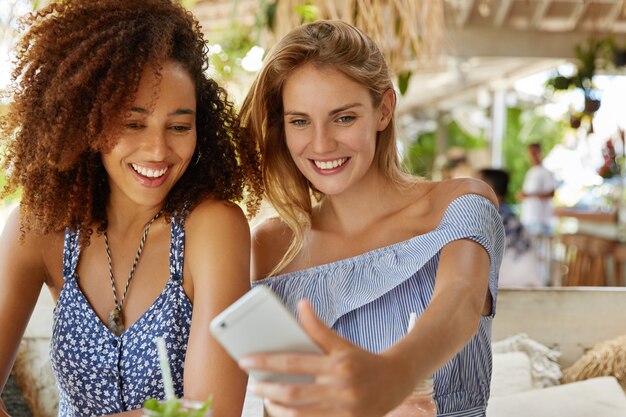 Encantadora joven afroamericana y su alegre novia posan juntas para selfie, usan un teléfono celular moderno, están contentas de encontrarse en la cafetería, tienen relaciones interraciales Tecnología y estilo de vida