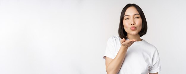 Encantadora joven asiática enviando besos al aire y mirando coqueta a la cámara de pie en camiseta sobre fondo blanco.