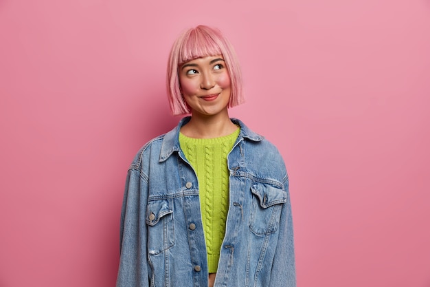 Encantadora joven asiática con expresión soñadora, humor romántico, mira en algún lugar arriba, viste un jersey verde de punto y una chaqueta vaquera, tiene un peinado bob,