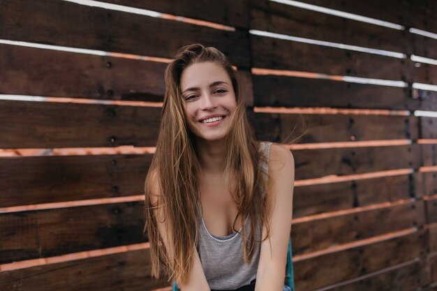 Encantadora joven blanca posando con placer en la pared de madera en primavera. Foto de niña alegre de pelo largo con hermosa sonrisa junto a la pared.