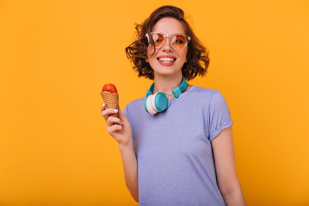 Encantadora joven comiendo helado durante la sesión de fotos Retrato interior de una guapa modelo femenina rizada que usa auriculares