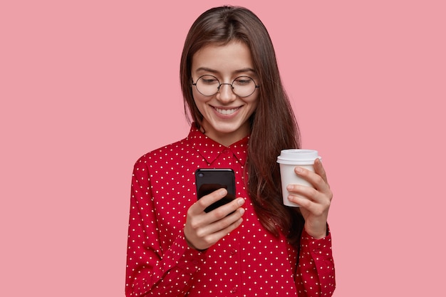 Encantadora joven complacida sostiene café caliente para llevar, teléfono móvil, feliz de recibir nuevo dispositivo como presente, viste camisa roja, tenía una sonrisa suave