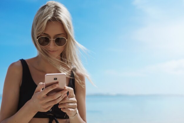 Encantadora joven disfrutando de las vacaciones de verano en la soleada playa de arena con móvil