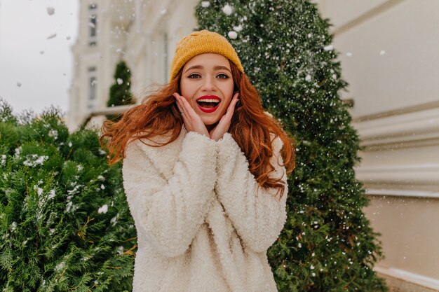 Encantadora joven jengibre posando en invierno. Chica bastante caucásica de pie junto a la picea con una sonrisa.