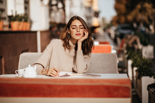 Encantadora joven morena con labios rojos, gafas y suéter beige, aprendiendo algo del cuaderno, tomando una taza de té en la terraza de un café en la cálida ciudad soleada