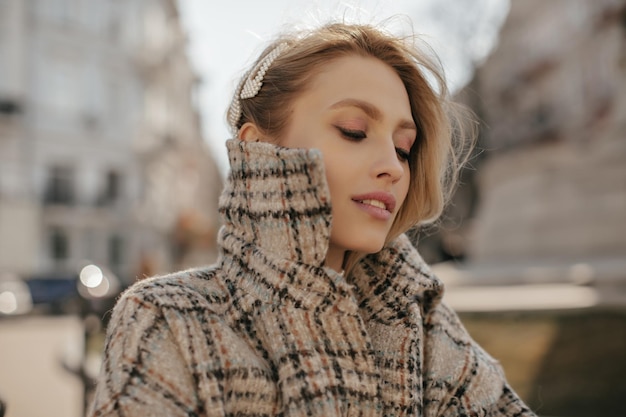 Encantadora joven mujer rubia con sonrisas de horquillas de perlas mira hacia afuera Bella dama en poses de abrigo cálido a cuadros en la plaza de la ciudad