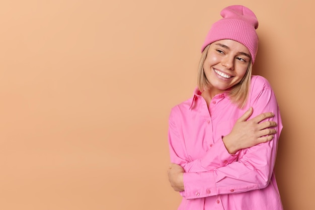 Encantadora joven soñadora con apariencia agradable se abraza a sí misma, sonríe felizmente, mira hacia otro lado, usa sombrero rosa y camisa aislada sobre fondo marrón, espacio de maqueta para su contenido publicitario