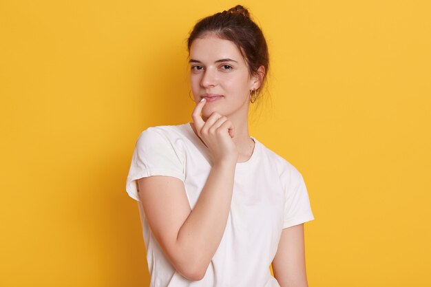 Encantadora joven vistiendo camiseta blanca, manteniendo el dedo en el labio