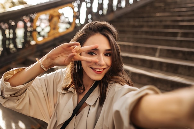 Encantadora jovencita con cabello castaño y labios rojos usando una gabardina beige sentada en las escaleras de la ciudad vieja y encantadora posando en la cámara al aire libre Cálido otoño soleado