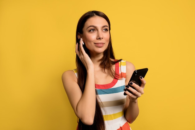 Encantadora linda chica con vestido de verano despojado escuchando música en auriculares y usando un teléfono inteligente sobre una pared amarilla
