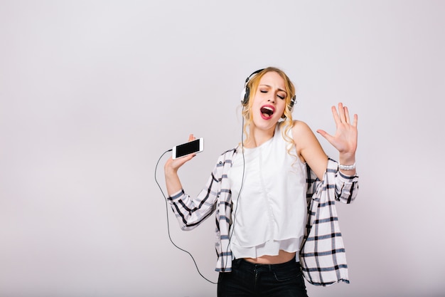 Encantadora linda mujer rubia disfrutando de su vida, cantando y bailando con los ojos cerrados. Chica inteligente alegre con elegante blusa blanca con pantalón negro.