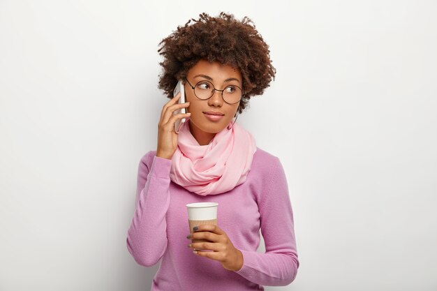 Encantadora linda mujer tranquila con piel oscura, peinado rizado, mira a un lado, hace una llamada a través de un teléfono inteligente, sostiene una taza de té desechable, viste un cuello alto de color púrpura con scrarf