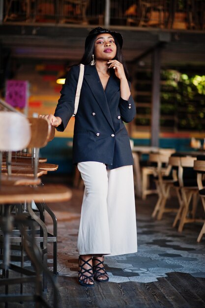 Encantadora modelo afroamericana con sombrero de chaqueta negra y riñonera relajándose en el café durante el tiempo libre