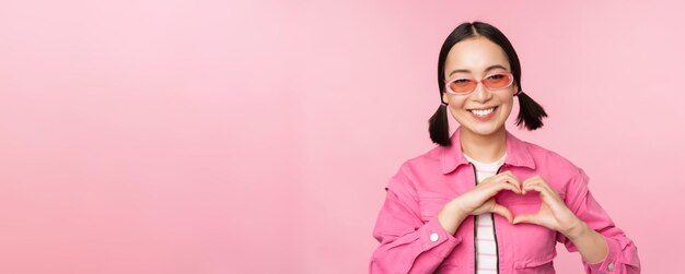 Encantadora modelo coreana con gafas de sol de moda muestra un signo de cuidado del corazón Te amo gesto sobre fondo rosa