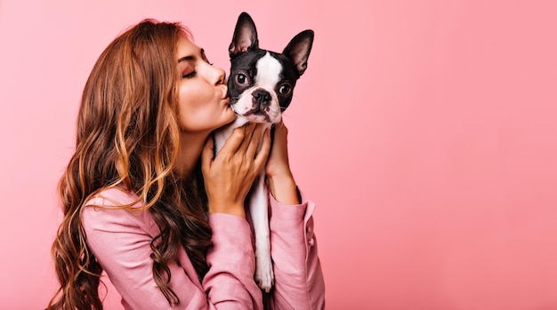 Encantadora modelo femenina besando a un divertido cachorro de bulldog Retrato interior de una refinada chica de pelo oscuro posando sobre fondo rosa con perro