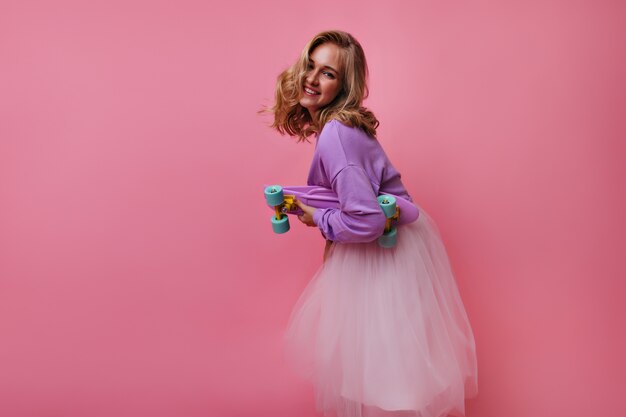 Encantadora modelo femenina en exuberante falda y camisa morada posando con patineta. Retrato de interior de chica rubia interesada juguetonamente