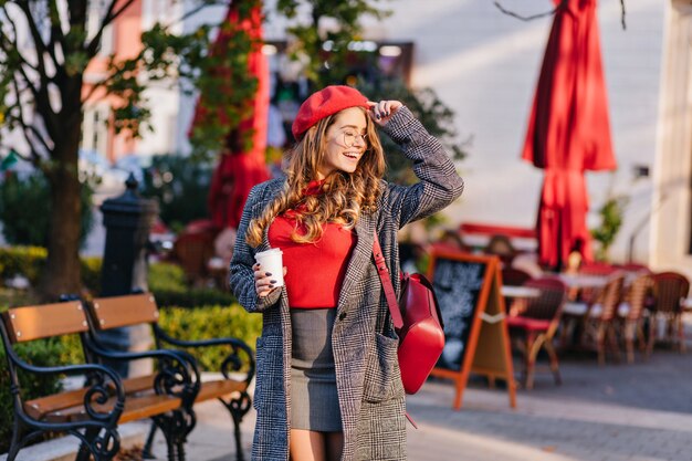 Encantadora modelo femenina en mini falda posando con los ojos cerrados en un día soleado en la calle cerca de la cafetería