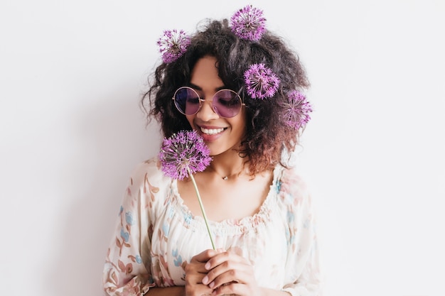 Foto gratuita encantadora modelo femenina con piel oscura sosteniendo allium y expresando felicidad. foto interior de mujer elegante y guapa con flor.