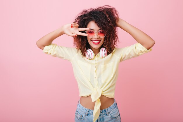 Encantadora modelo femenina en traje de verano disfrutando del tiempo libre y bailando. Retrato interior de maravillosa dama africana jugando con cabello oscuro y posando con el signo de la paz.