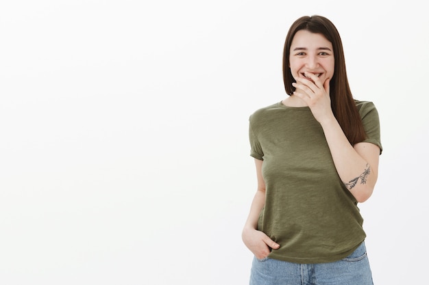 Encantadora morena de 20 años con tatuaje en el brazo riendo y sonriendo cubriendo la boca tímida mientras se ríen de escuchar una historia divertida, divirtiéndose hablando divertido y alegre sobre una pared gris