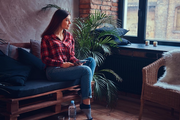 Encantadora morena con camisa de franela y jeans sentada en un sofá