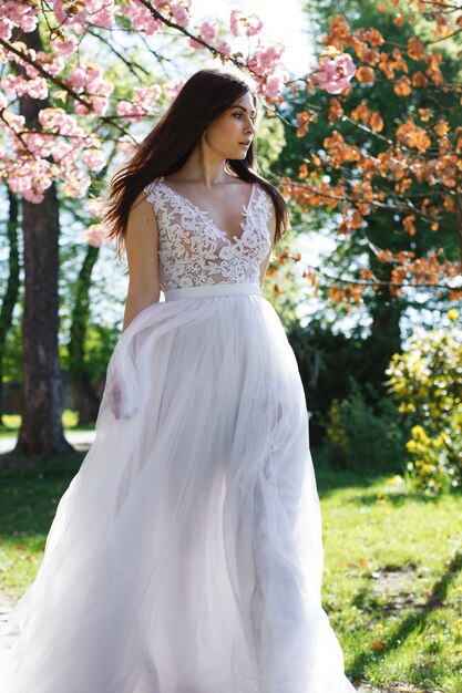 Encantadora morena novia camina en vestido blanco entre los árboles florecientes de sakura