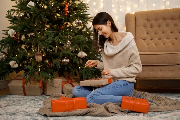 Encantadora mujer abriendo caja de regalo de Navidad, sentada en el piso cerca del árbol festivo en casa