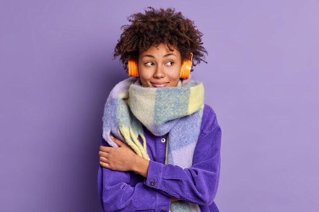 Encantadora mujer afroamericana pensativa se abraza a sí misma para abrigarse viste bufanda alrededor del cuello concentrada agradablemente a un lado mientras escucha música a través de auriculares inalámbricos.