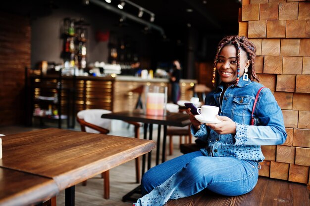 Encantadora mujer afroamericana con rastas en chaqueta de jeans con estilo azul en el café sostenga el teléfono móvil