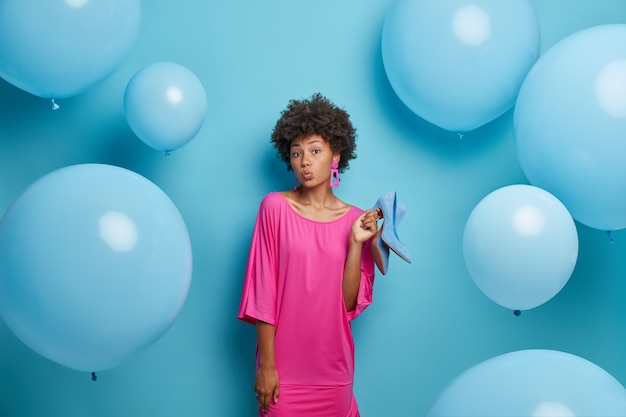 La encantadora mujer afroamericana rizada de piel oscura mantiene los labios redondeados, se viste con un vestido elegante, sostiene los zapatos, elige qué ponerse o qué vestirse, aislado en la pared azul con globos inflados alrededor.