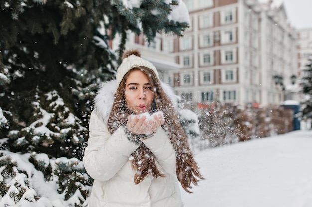 Encantadora mujer alegre que sopla copos de nieve de sus manos en día de invierno al aire libre en la calle.
