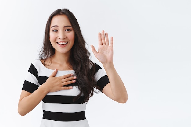 Encantadora mujer del este de Asia en camiseta a rayas haciendo promesa, levanta una mano y presiona la palma contra el corazón mientras jura decir la verdad, promete, tratando de ser honesto, sonriendo amigable y extrovertido, fondo blanco