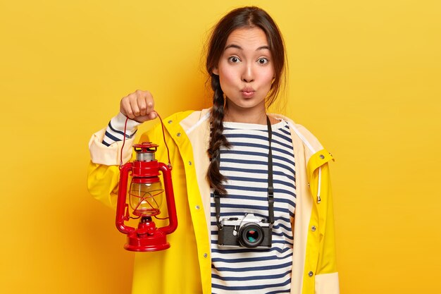 Encantadora mujer asiática con cabello largo y oscuro, sostiene una antorcha roja, vestida con un impermeable amarillo informal y un jersey a rayas, siendo un turista activo, caminatas durante el verano, captura el momento con una cámara retro