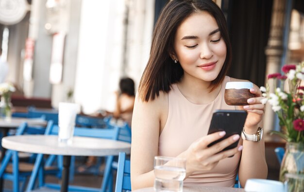 Encantadora mujer asiática con hermosa sonrisa leyendo buenas noticias en el teléfono móvil durante el descanso en la cafetería.