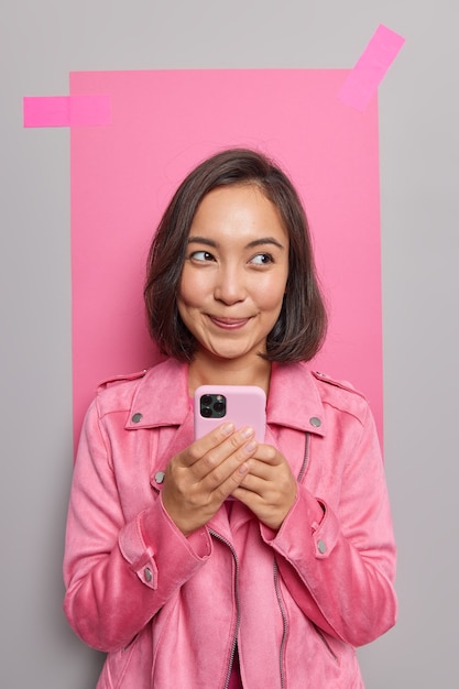 Encantadora mujer asiática joven y soñadora sostiene conversaciones de teléfono móvil con amigos, usa un gadget y una aplicación geniales vestida con poses de chaqueta contra la pared gris con papel rosa enlucido