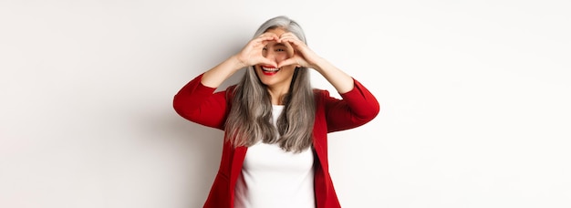 Foto gratuita encantadora mujer asiática de mediana edad con cabello gris con chaqueta roja que muestra el signo del corazón y mira a escondidas