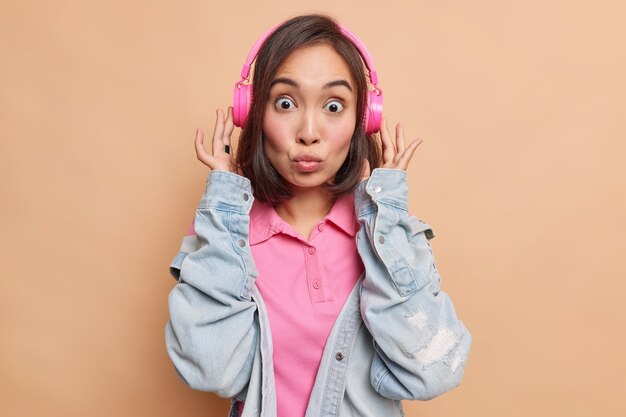 La encantadora mujer asiática morena con cabello oscuro mantiene los labios doblados tiene una expresión de asombro escucha música a través de auriculares inalámbricos rosas usa una chaqueta de mezclilla aislada sobre una pared beige