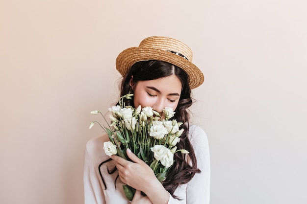 Encantadora mujer asiática oliendo flores blancas. Foto de estudio de mujer china con eustomas aislado sobre fondo beige.