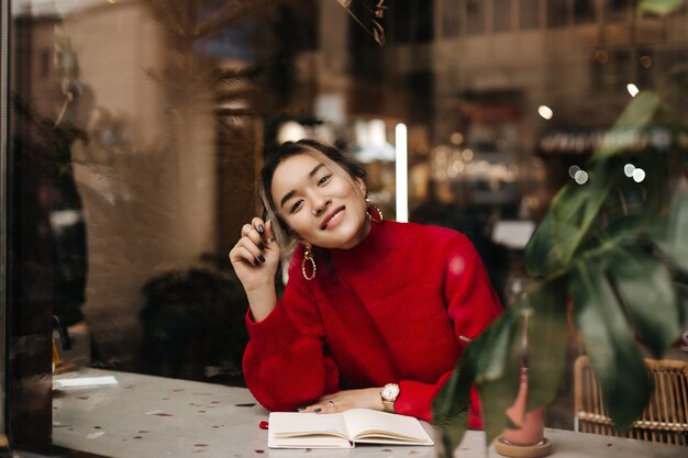 Encantadora mujer asiática en traje de punto rojo y enormes aretes sonríe mientras está sentado en la cafetería con el cuaderno en la mesa