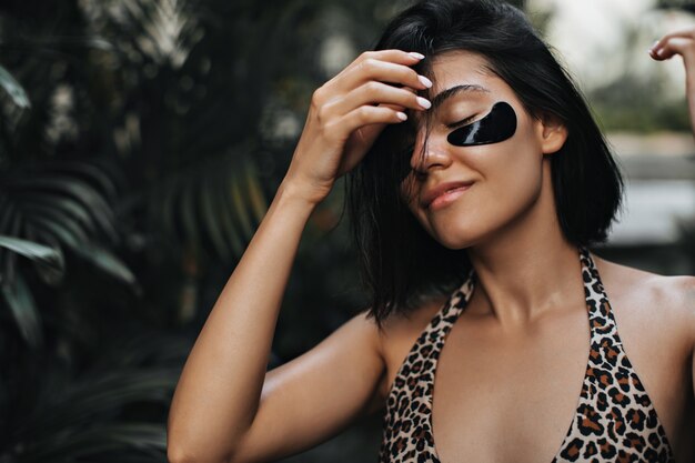 Encantadora mujer bronceada disfrutando de vacaciones en el resort. Mujer atractiva con parches en los ojos tocando el cabello sobre fondo de naturaleza.