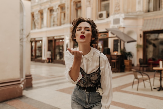 Encantadora mujer con cabello ondulado en camisa con encaje y jeans que sopla beso en la calle. Mujer morena con labios rojos posando en la ciudad.