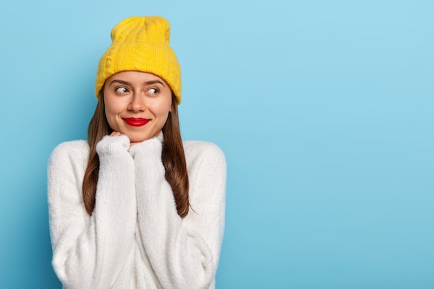 Encantadora mujer con cabello oscuro, lápiz labial rojo, mantiene las manos debajo de la barbilla, mira a un lado, imagina algo agradable