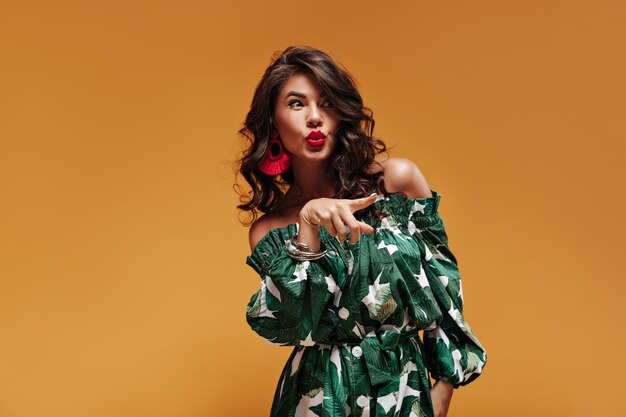 Encantadora mujer con cabello oscuro rizado y lápiz labial brillante en vestido verde con un gran botón blanco posando en un fondo aislado