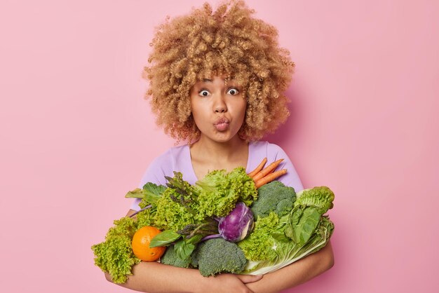 Encantadora mujer de cabello rizado mantiene los labios doblados sostiene un ramo de comestibles vegetales verdes frescos recogidos en el invernadero elige un estilo de vida de bienestar saludable aislado sobre fondo rosa Vitaminas de primavera