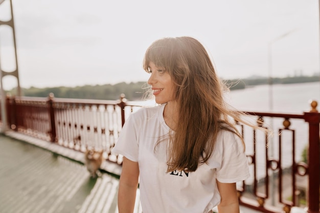 Encantadora mujer con camiseta blanca con cabello largo mira a un lado y sonríe mientras camina por el puente con un perro en la ciudad Chica de cabello oscuro con maquillaje desnudo posando a la luz del sol