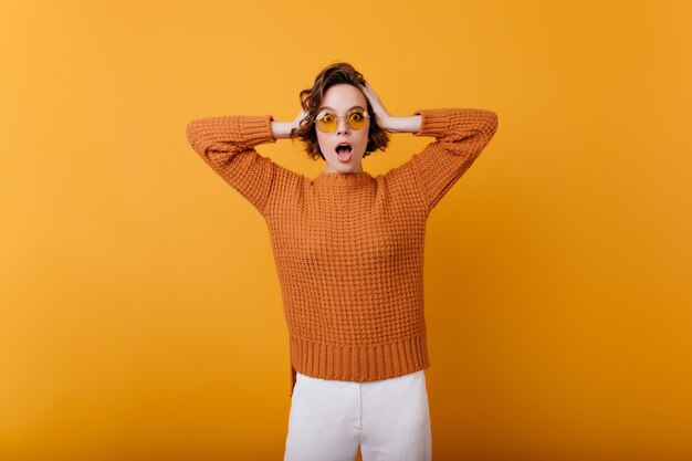 Encantadora mujer con cara de asombro epxression tocando su cabeza. chica europea emocional en suéter de lana de moda aislado en la pared naranja.