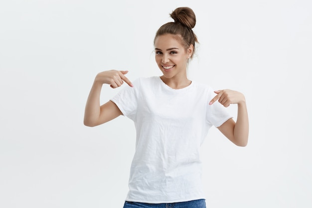 Encantadora mujer caucásica emotiva apuntando hacia abajo o hacia su camiseta mientras sonríe alegremente y expresa emociones positivas